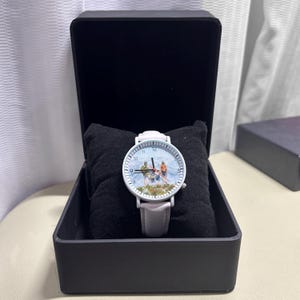 May include: A white wristwatch with a light gray leather band, displayed in a black box. The watch face features a mountain scene with skiers. The watch is resting on a black pillow.