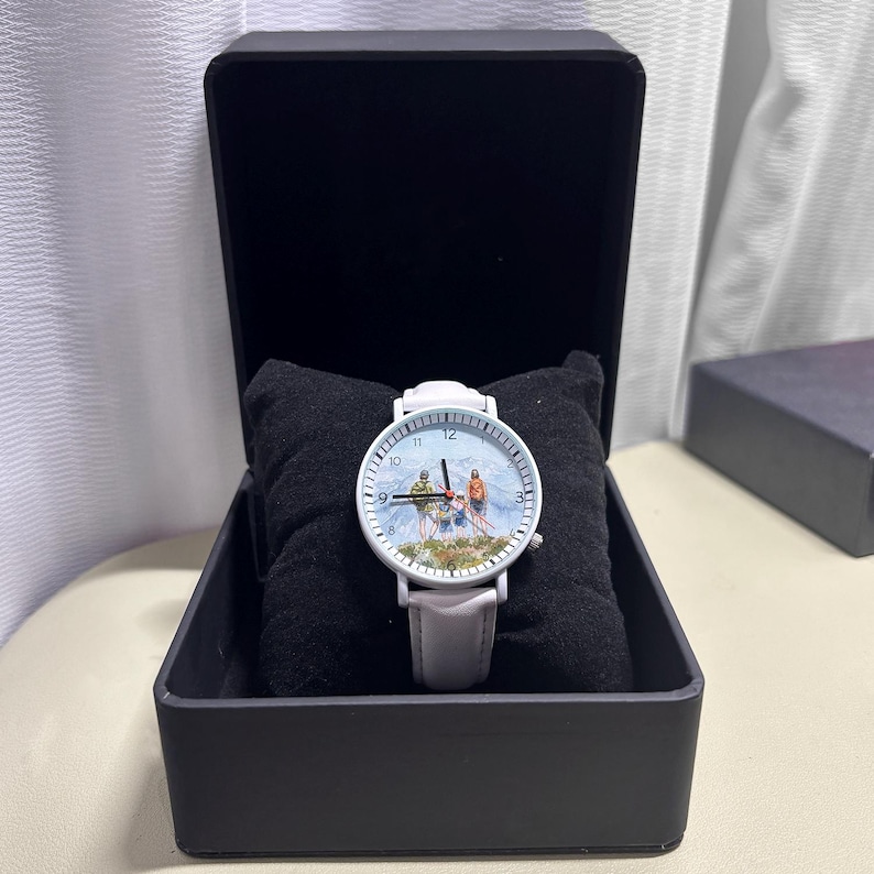 May include: A white wristwatch with a light gray leather band, displayed in a black box. The watch face features a mountain scene with skiers. The watch is resting on a black pillow.