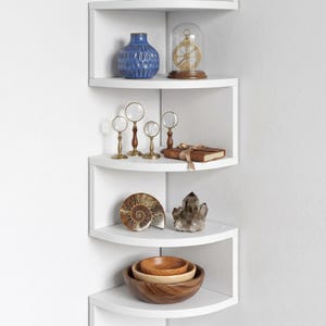 May include: A white corner shelf unit displaying decorative items. Includes a blue vase, a glass dome with a compass, magnifying glasses, a book, a shell, a crystal, and wooden bowls. The shelves are arranged in a zig-zag pattern.