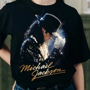 May include: Black t-shirt with a gold 'Michael Jackson' signature and a portrait of the singer in a hat and sequined jacket. The image shows a close-up of the artist in a performance pose.