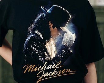 May include: Black t-shirt featuring a gold-colored Michael Jackson signature and a portrait of the artist wearing a hat and sequined jacket. The image is a close-up of the singer in a performance pose.