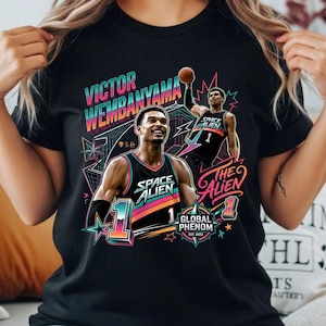 Könnte beinhalten: Schwarzes T-Shirt mit einer farbenfrohen Grafik eines Basketballspielers, dem Text "VICTOR WEMBANYAMA" und "SPACE ALIEN". Das Design enthält die Wörter "THE ALIEN" und "GLOBAL PHENOM EST. 2023".