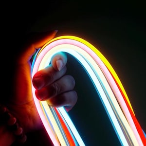 May include: A hand grips a flexible LED light strip, showcasing a spectrum of colours. The strip features parallel tubes in yellow, pink, white, blue, and red. The dark background accentuates the vibrant illumination of the light strip.
