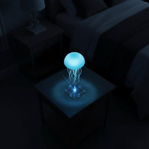 Könnte beinhalten: Eine quallenförmige Lampe mit blauem Leuchten. Die Lampe hat einen abgerundeten Kopf und fließende, tentakelartige Stränge. Sie steht auf einem transparenten, quadratischen Sockel und ist eingesteckt. Die Lampe steht auf einem dunklen Nachttisch.