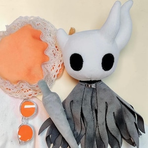 Könnte beinhalten: Ein Plüschtier einer Figur mit weißem Kopf, großen schwarzen Augen und einem grauen Umhang. Das Spielzeug hat ein kleines, graues Schwert und befindet sich neben einem orangefarbenen Plüsch mit weißer Spitzenborte.
