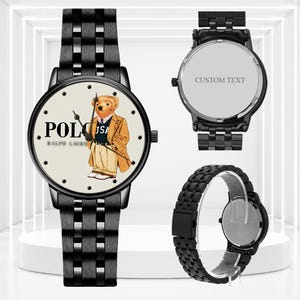 May include: A black metal wristwatch featuring a cream-colored dial with a graphic of a teddy bear wearing a tan coat and a navy sweater. The watch has a black link band. The back of the watch case is silver and includes the text CUSTOM TEXT.