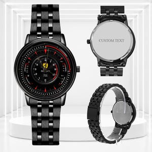 Pode incluir: Um relógio de pulso de aço inoxidável preto com mostrador redondo estilo tacômetro, detalhes em vermelho, um logotipo da Ferrari e o texto RPM x1000. A parte traseira da caixa é prateada e apresenta o texto CUSTOM TEXT.