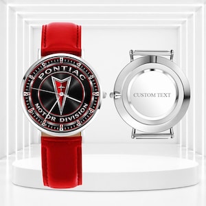 Könnte beinhalten: Eine Armbanduhr mit einem leuchtend roten Lederarmband und einem runden Zifferblatt, das das schwarz-rote Pontiac Motor Division Logo und silberne Zeiger zeigt. Daneben liegt ein silbernes Uhrengehäuse mit der Gravur CUSTOM TEXT in der Mitte.