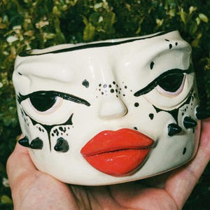 Puede incluir: Maceta de cerámica color crema con diseño de rostro estilizado. Presenta delineador negro, cejas y pecas, con púas negras a cada lado. La maceta tiene labios de color rojo brillante.