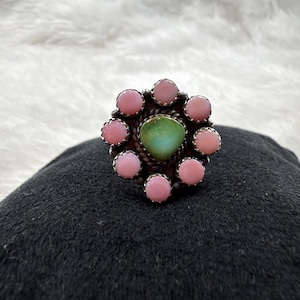 Puede incluir: Un anillo de plata con una piedra verde central en forma de lágrima rodeada por ocho piedras rosas redondas y lisas. El engaste incluye un detalle de cuerda metálica trenzada alrededor de la piedra central, con cada piedra rosa en un bisel.