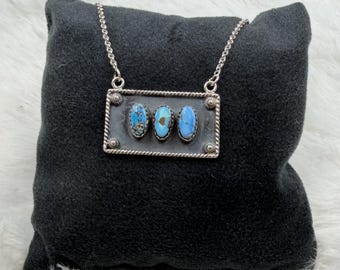 May include: A silver-toned rectangular pendant necklace featuring three oval-shaped blue turquoise stones set horizontally in a row. The pendant has a twisted rope-style border and is suspended from a delicate silver-toned chain link necklace.