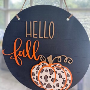 May include: A round wooden sign with a black background and the words "Hello Fall" in orange and gold lettering. A leopard print pumpkin is centered on the sign.