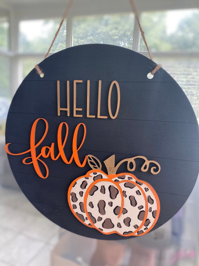 May include: A round wooden sign with a black background and the words "Hello Fall" in orange and gold lettering. A leopard print pumpkin is centered on the sign.