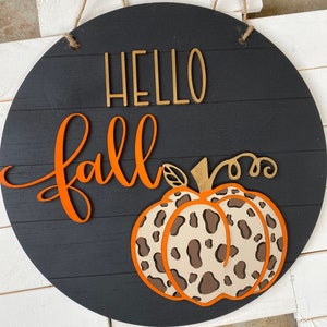 May include: Black wooden sign with the words "Hello Fall" in orange and gold lettering. A brown and white leopard print pumpkin is in the bottom center of the sign.