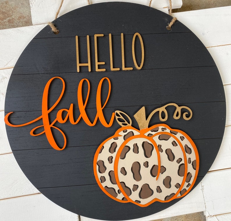 May include: Black wooden sign with the words "Hello Fall" in orange and gold lettering. A brown and white leopard print pumpkin is in the bottom center of the sign.