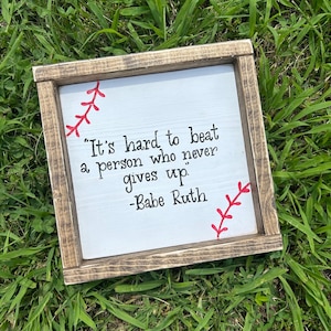 May include: A wooden sign with a white background and black text. The sign features a baseball stitch design in red and the quote "It's hard to beat a person who never gives up." - Babe Ruth.