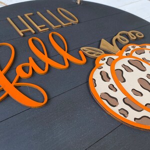 May include: A round wooden sign with the words "Hello Fall" in gold and orange lettering. A brown and white leopard print pumpkin is on the right side of the sign.