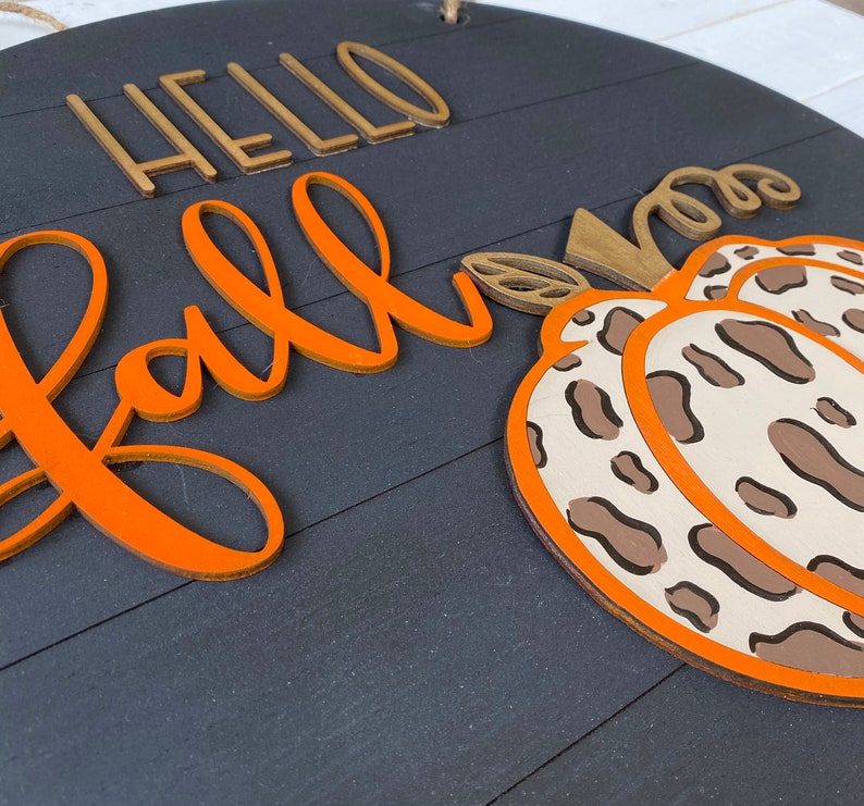 May include: A round wooden sign with the words "Hello Fall" in gold and orange lettering. A brown and white leopard print pumpkin is on the right side of the sign.