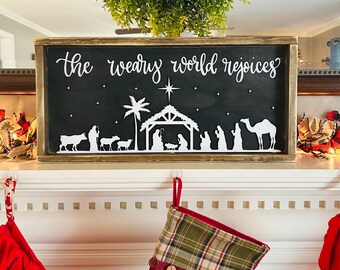 May include: A black wooden sign with white lettering that reads "The weary world rejoices." The sign features a silhouette of the nativity scene with a star above the stable. The sign is hanging on a white fireplace mantel with red and green stockings hanging on hooks.