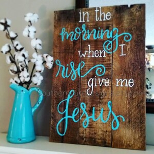 May include: Wooden sign with the text "in the morning when I rise give me Jesus" painted in turquoise blue on a brown wooden background.