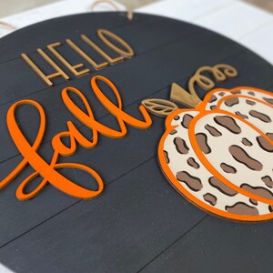 May include: A round wooden sign with the words "Hello Fall" in orange and gold lettering. A brown and white leopard print pumpkin is on the right side of the sign.