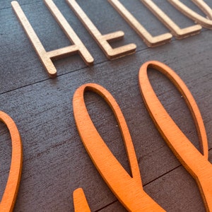 May include: A wooden sign with the words "Hello Fall" in gold and orange paint. The sign has a rustic wood grain texture.