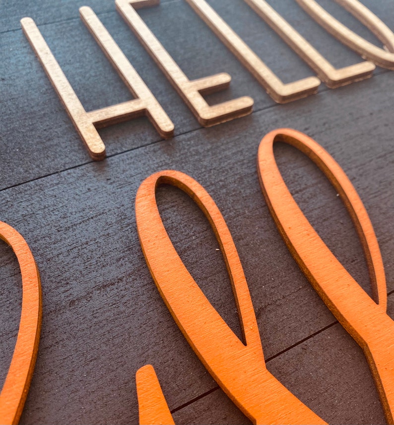 May include: A wooden sign with the words "Hello Fall" in gold and orange paint. The sign has a rustic wood grain texture.