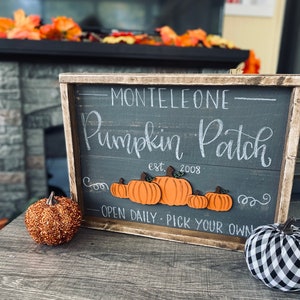 May include: A wooden sign with the words "Monteleone Pumpkin Patch" in white script, featuring orange pumpkin cutouts. The sign is framed in a rustic wooden border. A glittery orange pumpkin and a black and white checkered pumpkin are on the table.