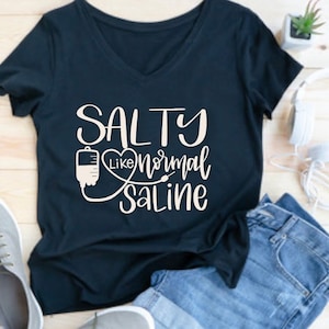 May include: A dark blue v-neck t-shirt with the text "SALTY like normal SALINE" in white. The text is in a handwritten font and includes a heart and an IV drip graphic.