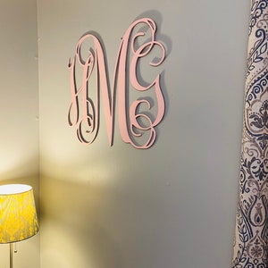 May include: A large, pink wooden monogram, featuring the letters "MHC" in a decorative script, is mounted on a gray wall. The monogram is the focal point, with a lamp and patterned curtain visible in the background.