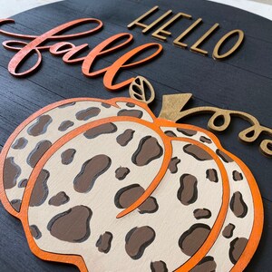 May include: A wooden sign with the words "Hello Fall" in gold letters and a large orange pumpkin with a brown and white leopard print design.
