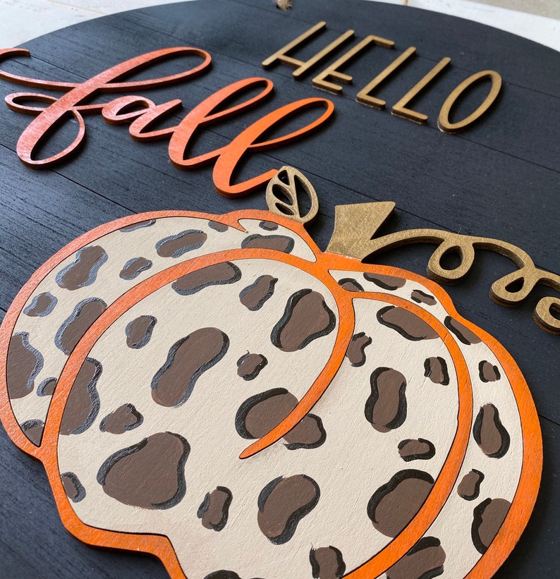 May include: A wooden sign with the words "Hello Fall" in gold letters and a large orange pumpkin with a brown and white leopard print design.