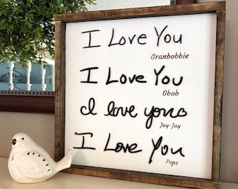 Op de afbeelding: Een wit houten bord met een bruin frame, waarop de zin "I Love You" vier keer in zwarte cursieve letters staat, waarbij elke inscriptie aan een andere persoon wordt toegeschreven: Granbobbie, Gbob, Joy-Joy en Pops.
