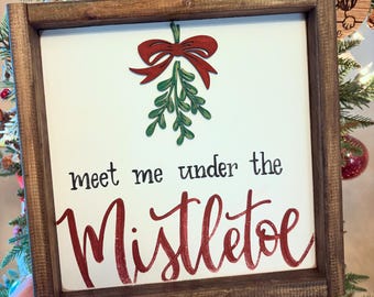 May include: A square wooden sign with a dark brown frame and a white background. The sign features a red bow and green mistletoe graphic at the top, with the black text meet me under the and the large red script text Mistletoe below.