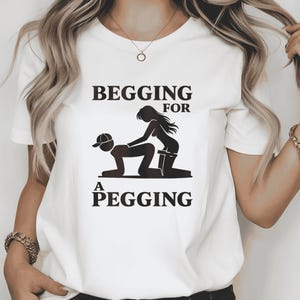Könnte beinhalten: Ein weißes kurzärmeliges T-Shirt mit einem schwarzen Grafikdruck von zwei Figuren in einer sexuellen Position. Der Text "BEGGING FOR A PEGGING" ist in schwarzen Großbuchstaben über und unter der grafischen Abbildung auf der Vorderseite gedruckt.