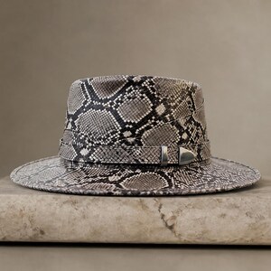 Puede incluir: Un sombrero fedora con un estampado de piel de serpiente en tonos grises y blancos. El sombrero tiene una corona estructurada, un ala ancha y una banda con una hebilla plateada. El sombrero se exhibe sobre un bloque de piedra.