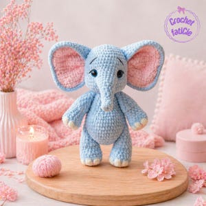 Peut inclure: Un éléphant en crochet bleu clair avec de grandes oreilles roses et une longue trompe. L'éléphant a des yeux bleus et se tient sur une surface en bois. L'image comprend le texte "Crochet faticle".