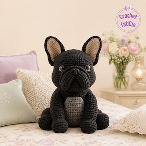 Puede incluir: Un perro Bulldog Francés negro tejido a mano con orejas color canela y ojos marrones. El perro tiene una mancha gris en el pecho y está sentado en una colcha con estampado floral. La imagen incluye el texto "Crochet faticie".