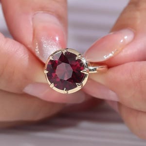Puede incluir: Un anillo con una gran piedra preciosa redonda de color rojo intenso engastada en una banda de oro pulido. La piedra está asegurada por múltiples garras que se extienden sobre el borde de la gema, creando un engaste firme y brillante.