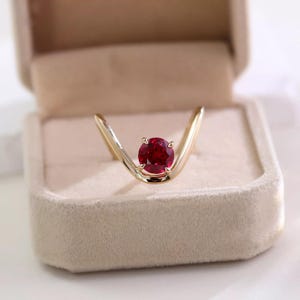 Pode incluir: Um anel de ouro em forma de V com uma única pedra preciosa redonda de cor vermelho profundo, fixada com quatro garras no centro da banda. A banda de ouro polido curva-se para cima em forma de chevron, destacando a pedra vermelha vibrante.