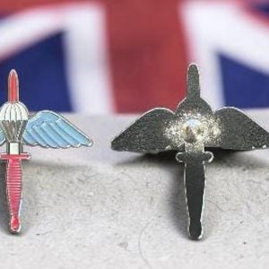 May include: Two metal lapel pins featuring a parachute and dagger design. One pin shows a red dagger, white parachute, and light blue wings. The other pin is a plain silver-toned metal version of the same design, accompanied by a small circular pin back.