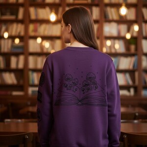 Puede incluir: Una sudadera morada de manga larga con un gráfico de líneas negras en la espalda. El diseño muestra un libro abierto con varias setas creciendo de las páginas. Otros pequeños diseños gráficos circulares son visibles en la manga derecha.