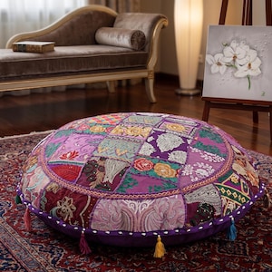 May include: A round floor cushion featuring a patchwork design of various purple, pink, and gold fabrics. The surface is decorated with intricate embroidery, floral patterns, and metallic thread accents, finished with small colorful tassels along the edge.