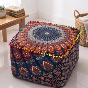 Puede incluir: Un otomán cuadrado con un vibrante diseño de mandala en tonos azules, naranjas y turquesas. La parte superior presenta un patrón circular detallado, y los bordes están adornados con pompones coloridos. Los lados muestran un patrón de cachemira y floral.