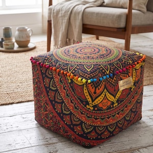 Könnte beinhalten: Quadratischer Hocker mit einem lebendigen Mandala-Design in Rot-, Orange- und Gelbtönen. Die Kanten sind mit bunten Pompons verziert. Der Hocker ist handbedruckt und steht auf einem hellen Holzboden.