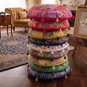 Puede incluir: Una pila vertical de nueve cojines de suelo redondos con diversos patrones de patchwork y colores vibrantes que incluyen rosa, rojo, dorado, negro, blanco, marrón, verde lima, verde esmeralda y azul, cada uno con pequeñas borlas decorativas.
