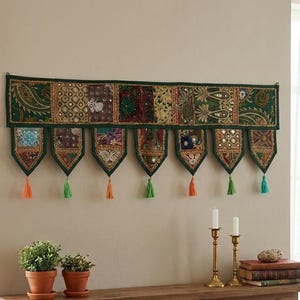 Op de afbeelding: Een rechthoekige wandhanger met een patchwork van donkergroene stoffen panelen versierd met ingewikkeld goudborduurwerk, lovertjes en spiegels. Zeven puntige stoffen hangers hangen aan de onderrand, elk afgewerkt met een kleurrijke kwast.