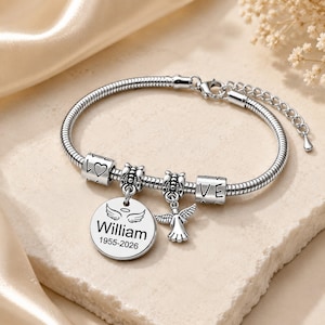 May include: A silver charm bracelet with a snake chain. The bracelet has charms spelling "LOVE" and "VE", plus a round charm engraved with "William" and the years "1955-2026", and an angel charm.