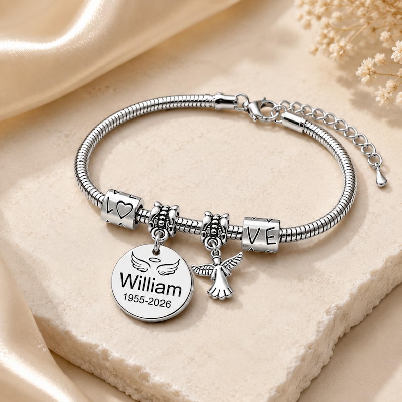 May include: A silver charm bracelet with a snake chain. The bracelet has charms spelling "LOVE" and "VE", plus a round charm engraved with "William" and the years "1955-2026", and an angel charm.