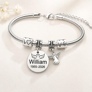 May include: A silver bracelet with several charms. The bracelet features a round charm engraved with the name "William" and the years "1955-2026". Other charms include a heart, the word "LOVE", and an angel. The bracelet is displayed on a white background.